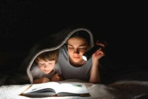 Annesi ile karanlıkta battaniyenin altında kitap okuyan çocuk